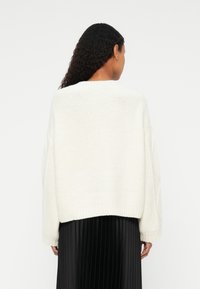 Suéter blanco de punto con un ajuste holgado, con hombros caídos y puños de canalé, combinado con una falda negra plisada.