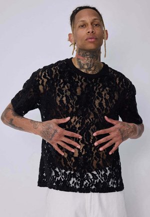 Personne avec des tatouages porte un haut court en dentelle fleurie noire et un pantalon blanc, posant les mains près de la taille sur un fond uni.