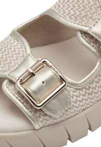 Chaussure tissée beige avec des accents métalliques, dotée d'une fermeture à boucle carrée et d'une semelle blanche épaisse et texturée.
