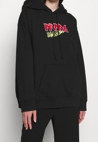 Svart hoodie i bomull med en frampocket, prydd med fet röd och gul broderad text på bröstet och snoddar på huvan.