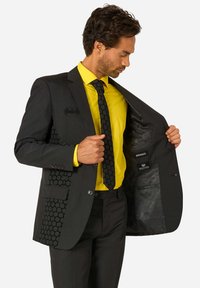 Mann in schwarzem Anzug mit Hexagonmuster und Fledermaus-Logo, gelbem Hemd und schwarzer Krawatte öffnet seine Jacke, um die Aufnäher „Wayne Industries“ und „Opposuits“ im Inneren zu zeigen.