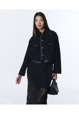 Jeune femme portant une veste en dentelle noire et une jupe assortie, tenant un sac à main noir texturé sur un fond blanc uni.