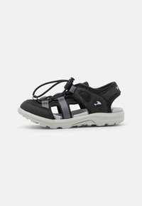 Sandal preto para exterior com parte superior em camurça e tiras elásticas cinzentas. Possui um fecho com cordão de puxar e uma sola branca grossa com aderência texturizada.