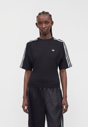 Personne aux cheveux tressés portant un t-shirt Adidas noir avec des bandes blanches sur les manches et un pantalon noir assorti sur un fond uni.