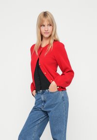 Red knit cardigan with round neckline, long sleeves, and gold buttons. Paired with a black top and high-waisted blue jeans. Smooth texture.