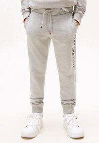 Osoba v svetlošedých teplákoch so stranovým logom a bielych teniskách, s rukami v vreckách, stojaca na bielom pozadí.