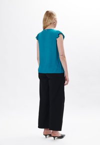 Top smanicato teal con spalle arricciate, tessuto testurizzato, abbinato a pantaloni neri a gamba larga e décolleté nere a punta.