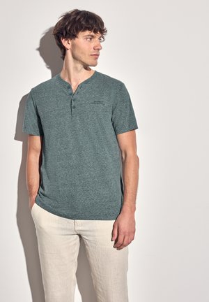 Camiseta de manga corta color verde jaspeado con cuello Henley, que presenta dos botones y un logo sutil, combinada con pantalones de color claro.