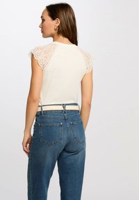 Maglietta beige chiaro con maniche a cappuccio in pizzo e scollatura rotonda, abbinata a jeans in denim blu a vita alta con tasche posteriori e cintura.