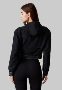 Czarna krótka kurtka z kapturem, fakturowana tkanina, elastyczne mankiety i pas, oraz dyskretna tylna kieszeń. Nosić na czarnych legginsach.