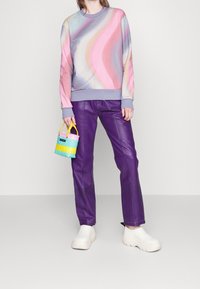 Sudadera multicolor con patrones pastel en espiral, pantalones de cuero morados, zapatos blancos grandes y un pequeño bolso de colorido tejido.