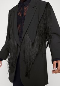 Blazer noir avec de longues franges noires aux épaules, design structuré, et un haut en dentelle noire transparente partiellement visible en dessous.