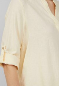 Chemise en lin jaune clair avec un col et des manches à boutons retroussées. Texture douce avec un léger brillant et des détails de couture minimaux.