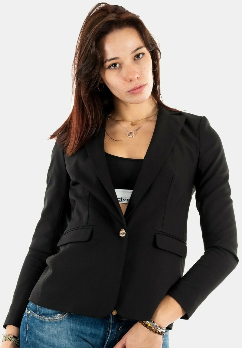 Blazer nero con una vestibilità aderente, un singolo bottone dorato, rever a aletto e due tasche frontali. Indossato sopra un top nero e jeans blu.