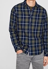 Camicia in flanella a quadri nei colori blu navy, verde e bianco; presenta un colletto button-down e maniche lunghe; abbinata a jeans grigi chiari.