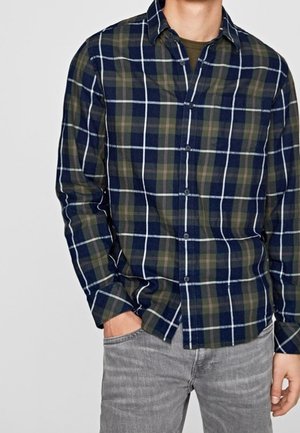 Karierte Flanellbluse in Marineblau, Grün und Weiß; mit Button-Down-Kragen und langen Ärmeln; kombiniert mit hellgrauen Jeans.
