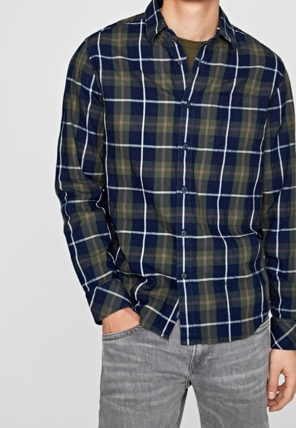 Camicia in flanella a quadri nei colori blu navy, verde e bianco; presenta un colletto button-down e maniche lunghe; abbinata a jeans grigi chiari.