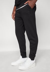 Svarte smale joggebukser med sidelommer og et teksturert logo. Kombinert med hvite treningssko.