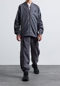 Mörkgrå idrottsträningsoveralljacka med dragkedja, ribbade ärmslut och logotyp på bröstet, i kombination med matchande cargobyxor och svarta sneakers.