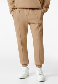 Beige Jogginghosen mit elastischen Bündchen und Seitennahtdetails, aus weichem Material, kombiniert mit weißen Turnschuhen.