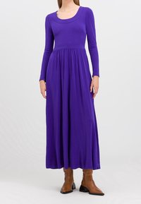 Lila långärmad maxiklänning i mjukt tyg med figursydd överdel och utsvängd kjol, med rundad halsringning och subtila draperingar vid midjan.
