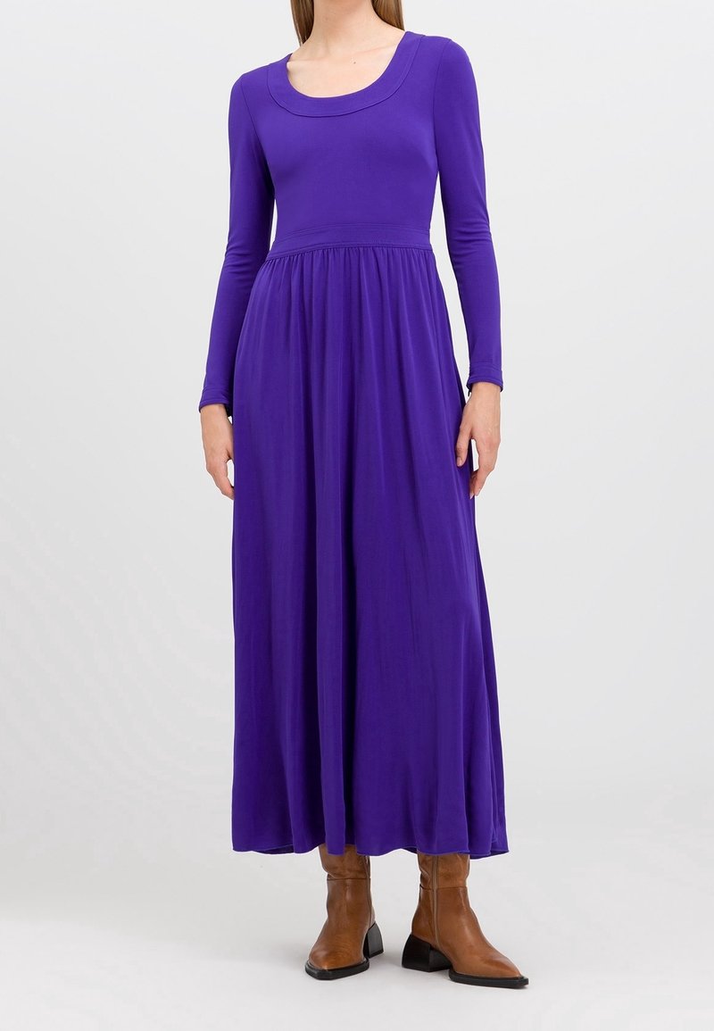 Lila långärmad maxiklänning i mjukt tyg med figursydd överdel och utsvängd kjol, med rundad halsringning och subtila draperingar vid midjan.