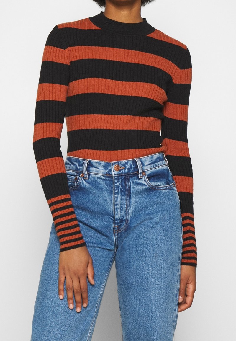 Personne portant un pull côtelé ajusté avec de larges rayures horizontales noires et orange et un jean bleu taille haute.