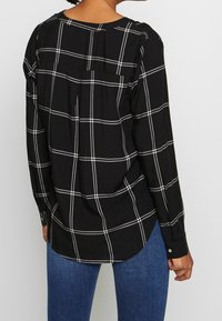 Chemise à carreaux noir et blanc à manches longues, décolleté rond, détails de boutons aux poignets et coupe décontractée. Fente au dos pour une meilleure mobilité.