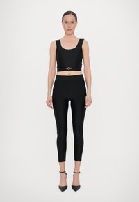 Top corto nero con spalline larghe e un dettaglio in metallo dorato, abbinato a leggings neri aderenti con una vita con logo, indossato con tacchi neri.