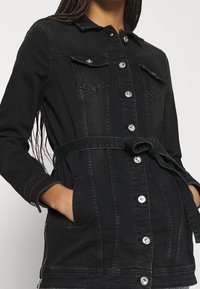 Černá džínová bunda s knoflíkovým zapínáním vpředu, dvěma náprsními kapsami, páskem v pase a dlouhými rukávy s mírným lemem. Hladká textura.
