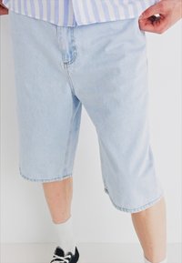 Light blue denim shorts with a loose fit, reaching mid-calf, featuring stitched hems and deep side pockets. Worn with white socks and black shoes.