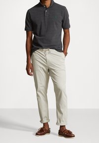 Mörkgrå pikétröja med korta ärmar, tre knappar och en grön logotyp. Kombinerad med ljusbeiga dragsko-byxor och bruna seglarskor.