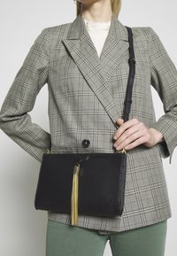 Musta nahkainen olkalaukku, jossa vetoketju ja kullanvärinen hapsukoriste. Laukulla on jämäkkä suorakulmainen muoto, ja se on puettu ruudullisen takin päälle.