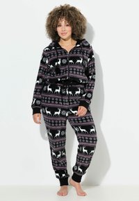 Mujer vistiendo un sudadera de forro polar negro y pantalones con patrones de renos y copos de nieve blancos, de pie descalza frente a un fondo blanco.