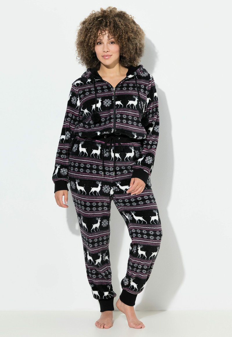 Mujer vistiendo un sudadera de forro polar negro y pantalones con patrones de renos y copos de nieve blancos, de pie descalza frente a un fondo blanco.