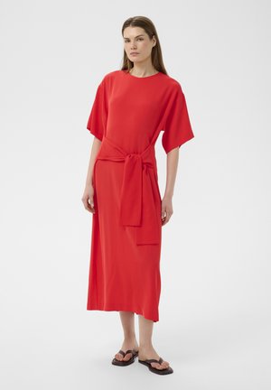 Femme debout dans une robe midi rouge à manches larges, avec un détail de taille noué, et des sandales plates noires sur un fond gris uni.