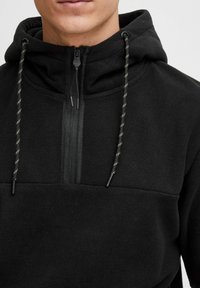 Schwarzer Fleece-Pullover mit hohem Kragen, halbem Reißverschluss und verstellbare Kapuze. Enthält kontrastierende, texturierte Kordeln mit gelben Akzenten.
