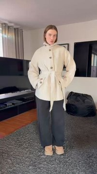 Kremo spalvos švarkas su diržu ir dideliais mygtukais, turintis stovimą apykaklę. Derinamas su juodomis plačiomis kelnėmis ir smėlio spalvos įmaukšlinamais batais.