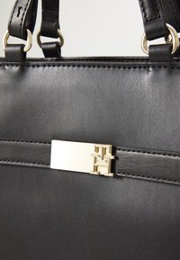 Sac à main en cuir noir avec un fermoir en métal de couleur dorée arborant un logo. Texture lisse avec des détails cousus et accents en métal.