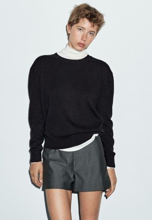 Czarny, lekko oversize sweter z długimi rękawami noszony na białej golfie, w połączeniu z szarymi szortami w kratę. Gładka faktura, minimalistyczny design.
