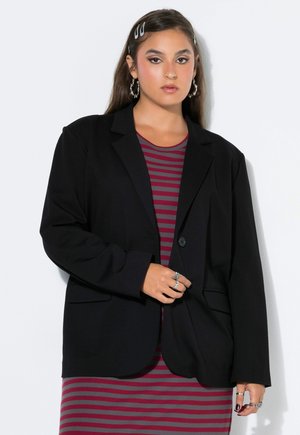 Mujer con cabello largo y castaño lleva una chaqueta negra sobre un vestido a rayas rojas y grises, complementado con pendientes de aro plateados, anillos y pasadores para el cabello.