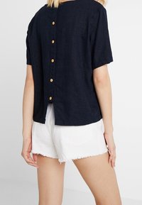 Tmavě modrá krátkosrstá košile s kulatým výstřihem, se řadou pěti dřevěných knoflíků vzadu a mírně otevřeným designem na zádech.