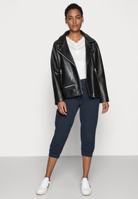 Chaqueta de motocicleta de cuero negro con cremalleras plateadas, superpuesta sobre una blusa blanca con encaje y combinada con pantalones cortos azul marino y zapatillas blancas.