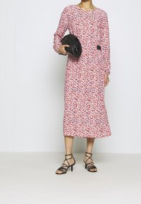 Vestito a maniche lunghe rosa con stampa floreale rossa e nera, aderente in vita, abbinato a una pochette nera e sandali neri con cinturino.