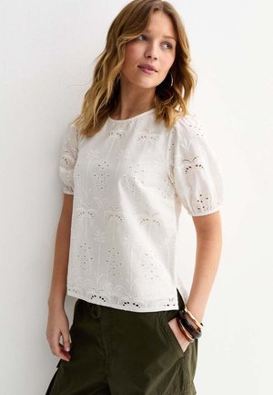 Kobieta o falowanych blond włosach, ubrane w białą koszulę z haftowanymi bufkami i oliwkowo zielone spodnie, z bransoletkami na lewej ręce.