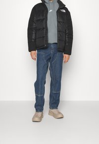 Persona vistiendo una chaqueta acolchada negra, sudadera gris de Nike, pantalones vaqueros azules con costuras reforzadas y zapatillas chunky beige, de pie contra una pared blanca lisa.