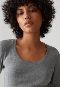 Woman with curly dark hair wearing a gray scoop-neck long-sleeve shirt against a plain light background.
