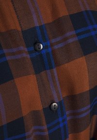 Gros plan d'un tissu à carreaux marron et bleu marine avec de fines lignes bleues et deux boutons noirs cousus.
