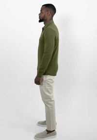 Man in olijfgroene langoor-polo, beige broek en lichtgrijze instappers staat in profiel op een witte achtergrond.