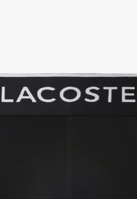 Zwarte boxershorts hebben een brede elastische tailleband met "LACOSTE" in witte letters geweven. De stof is glad en rekbaar.